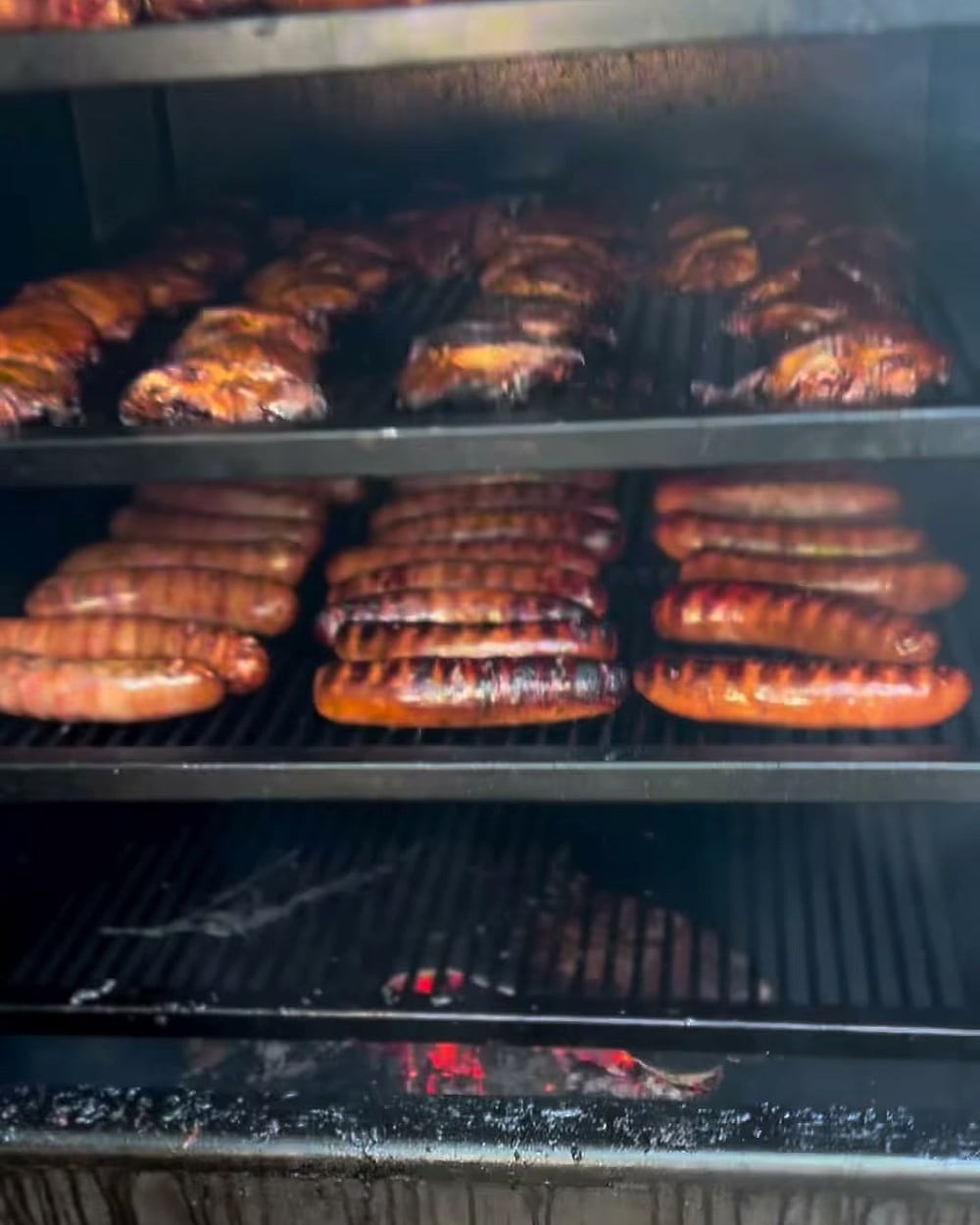 Quarts de poulet et saucisses alignés sur les grilles du smoker, braises visibles en dessous.